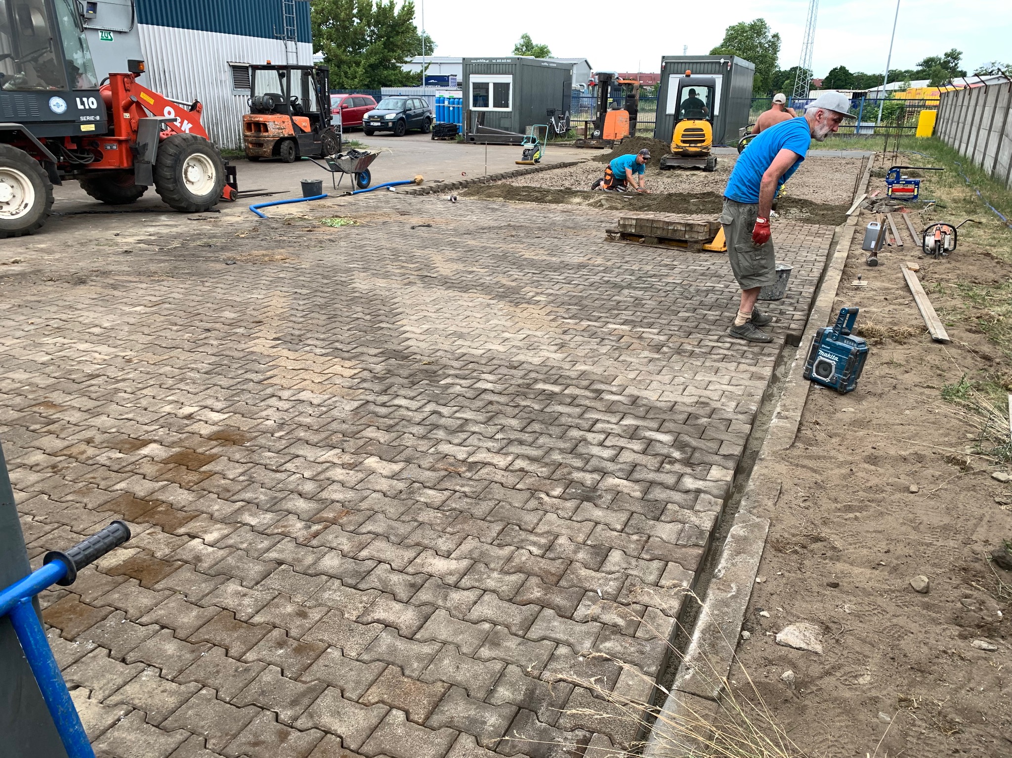 Prace brukarskie w toku: robotnicy układają kostkę, widoczne maszyny budowlane, w tym mini koparki i zagęszczarka. Częściowo ułożona nawierzchnia z kostki.