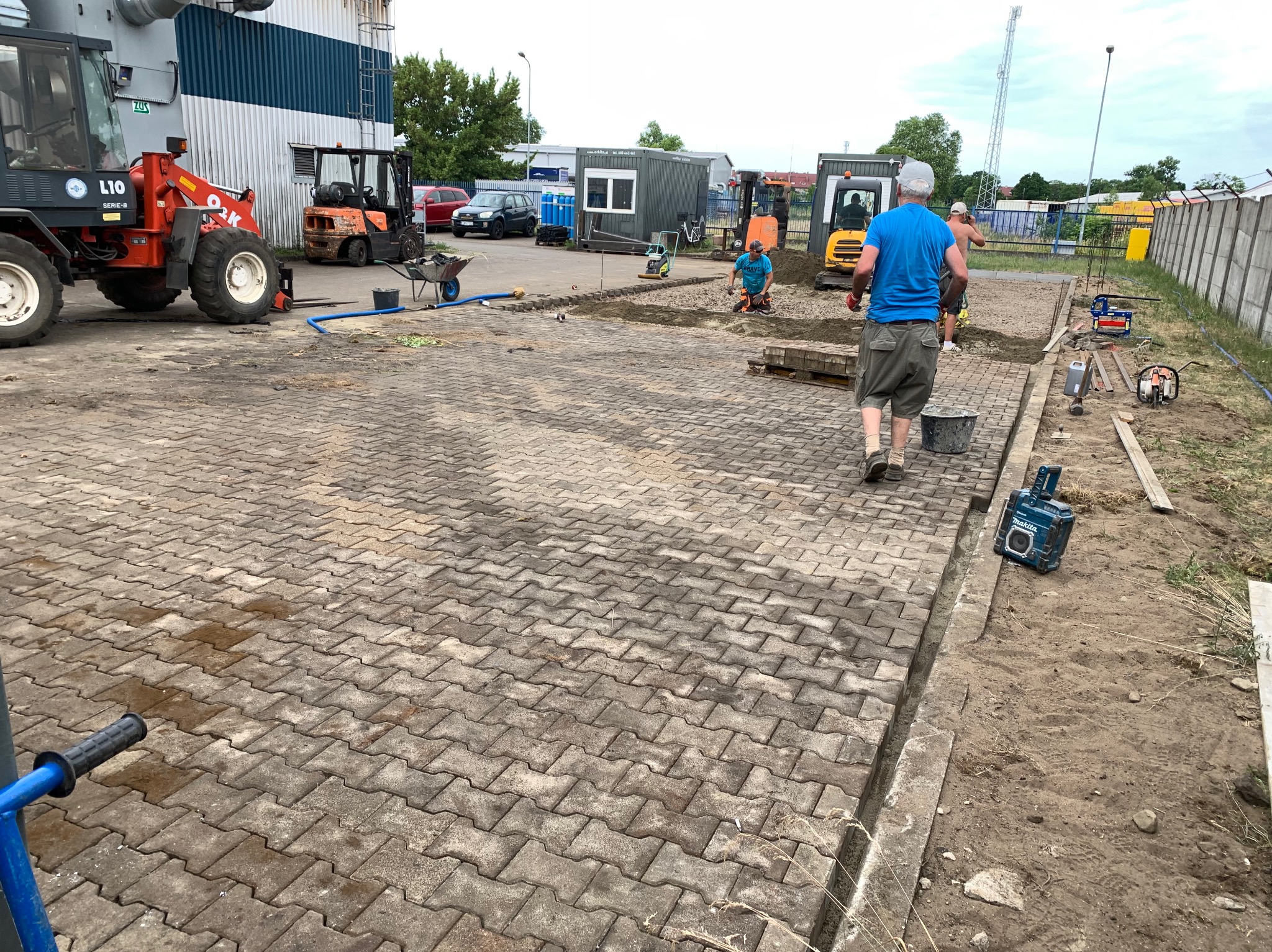 Prace brukarskie w toku: robotnicy układają kostkę na placu z użyciem mini-koparki i zagęszczarki. Część kostki już ułożona, widoczne narzędzia i materiały budowlane.