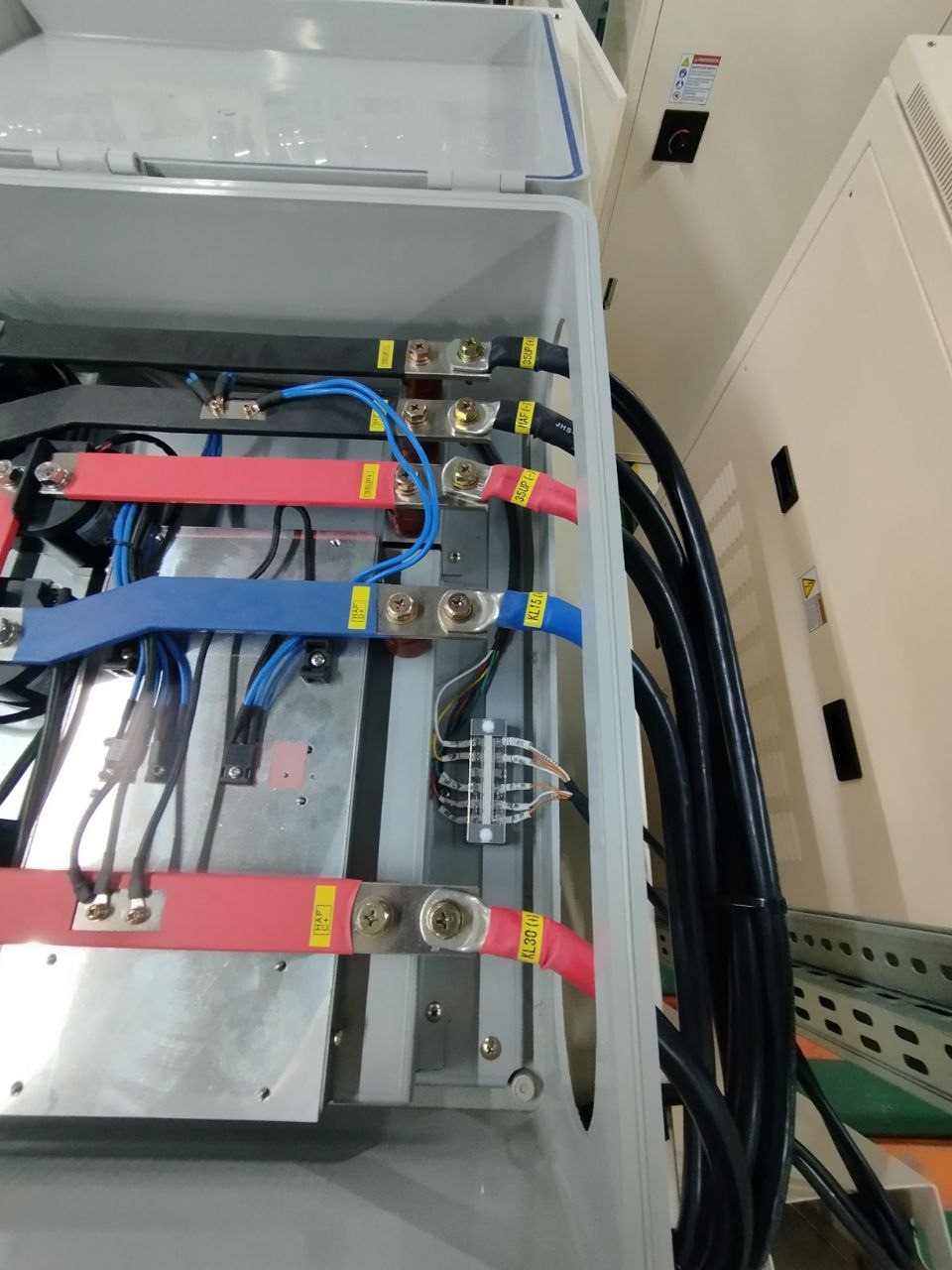 Wnętrze skrzynki elektrycznej z kolorowymi przewodami (czerwonymi, niebieskimi, czarnymi) podłączonymi do metalowych szyn za pomocą śrub. Widoczne kable i elementy montażowe.