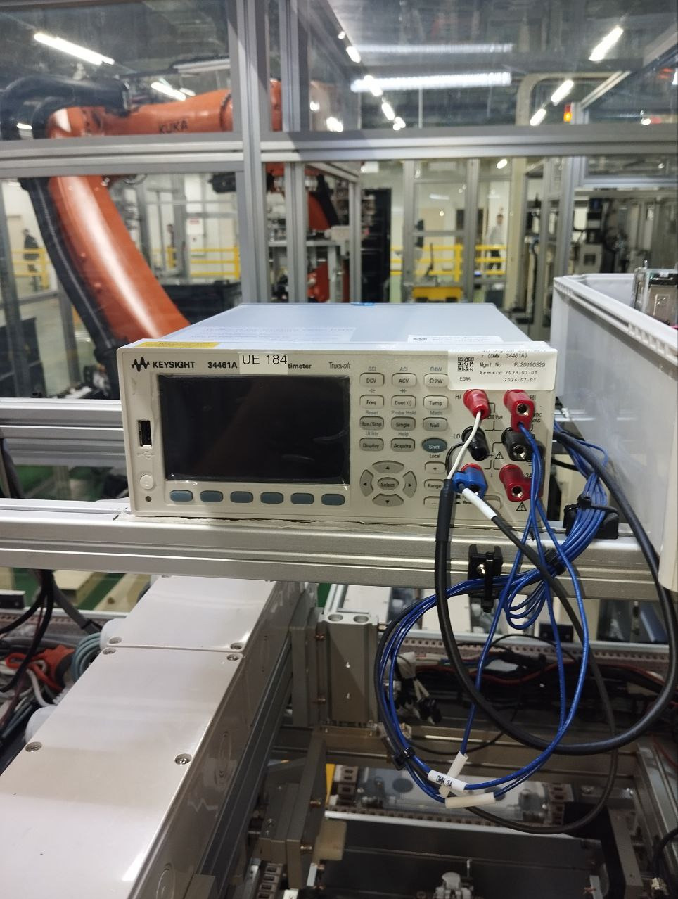 Multimetr Keysight UE 184 na stanowisku testowym z robotem KUKA w tle. Kable pomiarowe podłączone. Widoczne przyciski i wyświetlacz urządzenia. Precyzyjny pomiar w automatyce.