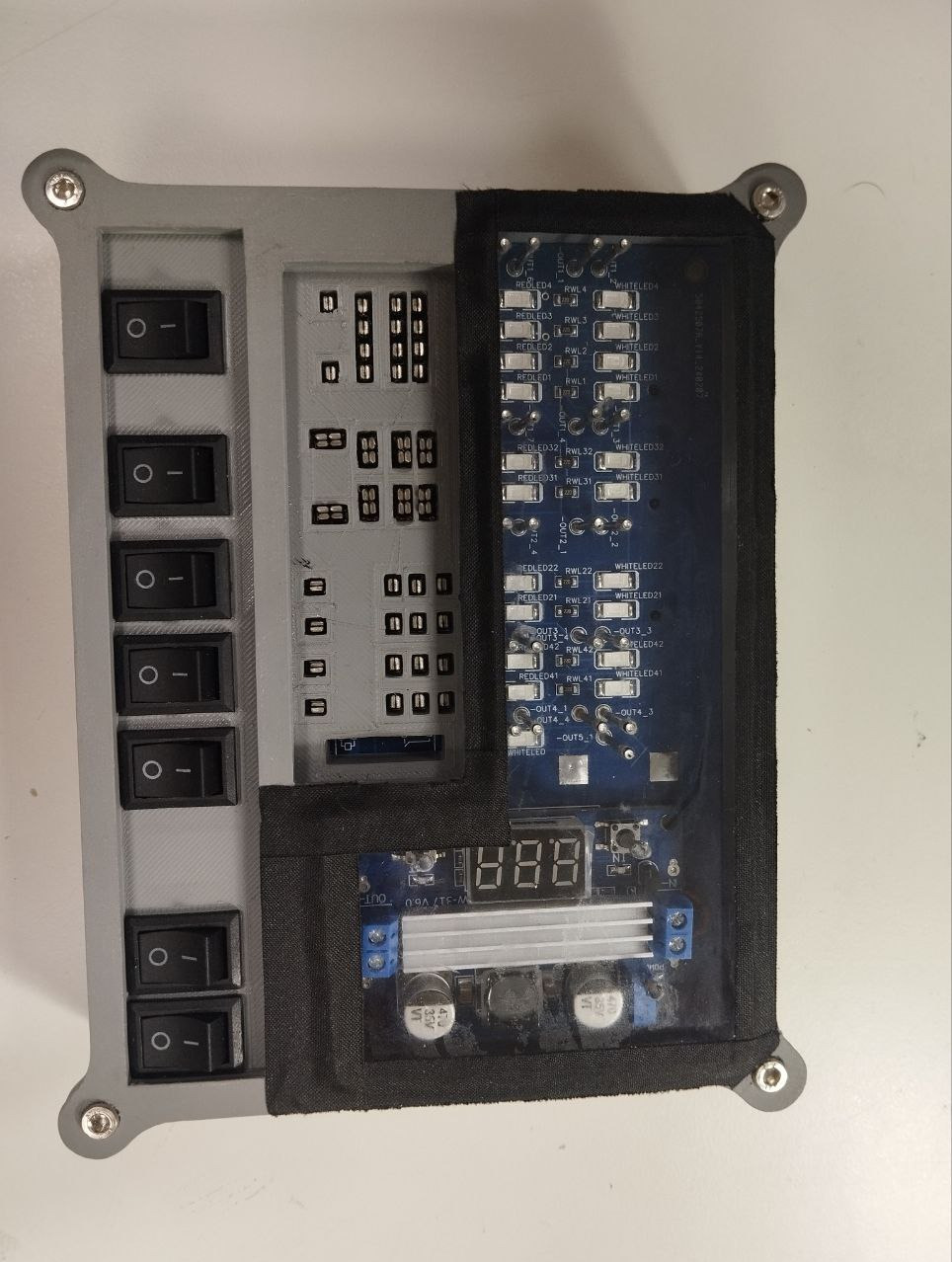 Szara obudowa z włącznikami, panelami sterowania i płytką drukowaną z diodami LED, potencjalnie element instalacji elektrycznej lub automatyki.