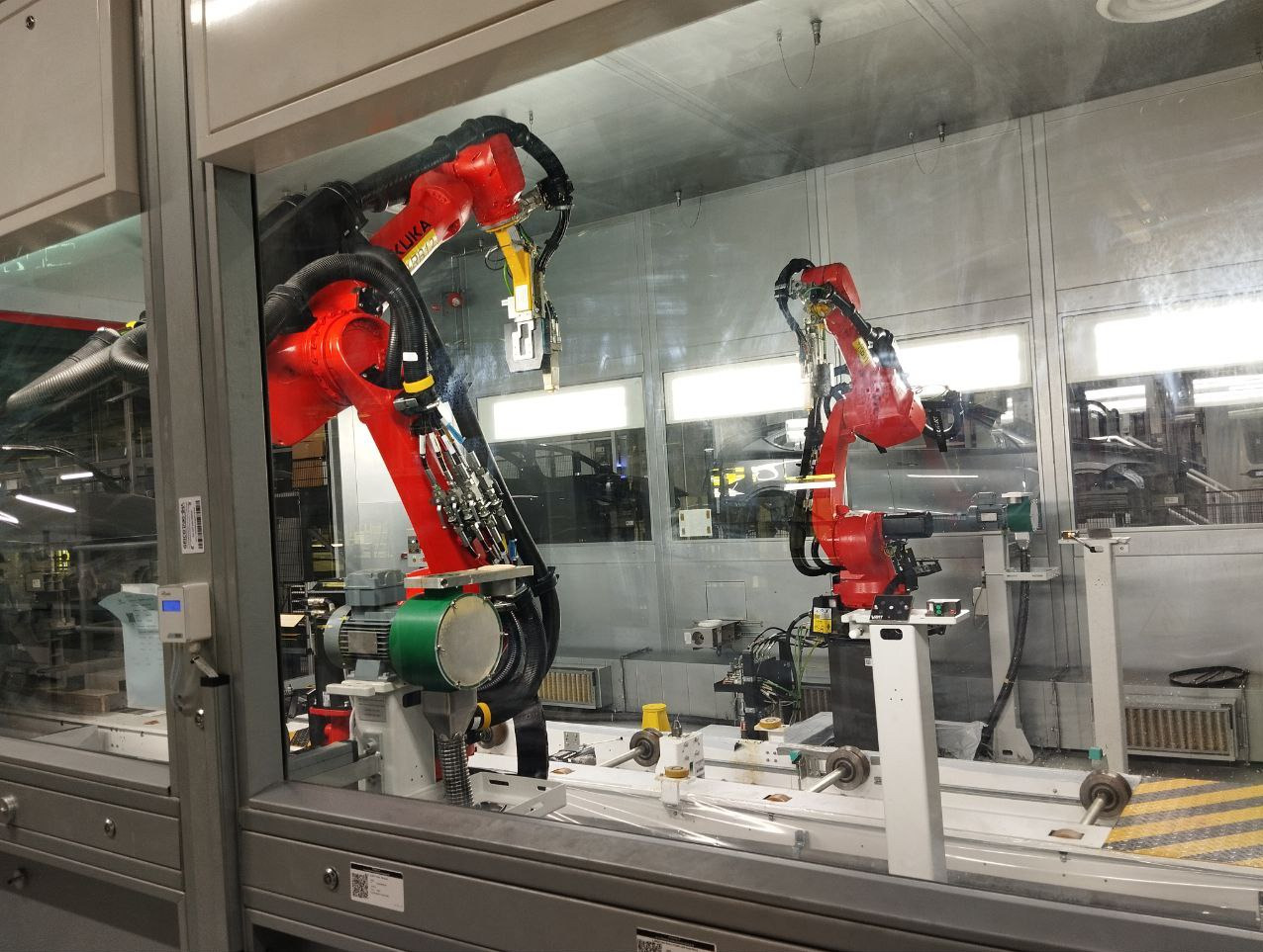 Dwa czerwone roboty przemysłowe KUKA pracujące w hali produkcyjnej za szklaną osłoną, automatyzacja procesów, precyzyjne ramię robota.