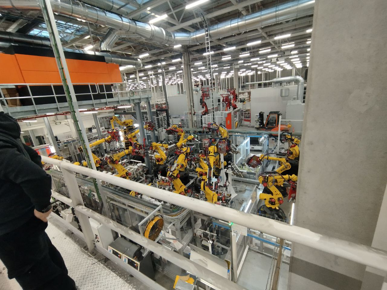 Rozległa hala fabryczna z licznymi żółtymi robotami przemysłowymi, widoczna z góry, z elementami konstrukcyjnymi i instalacjami pod sufitem. Lekkie oświetlenie.