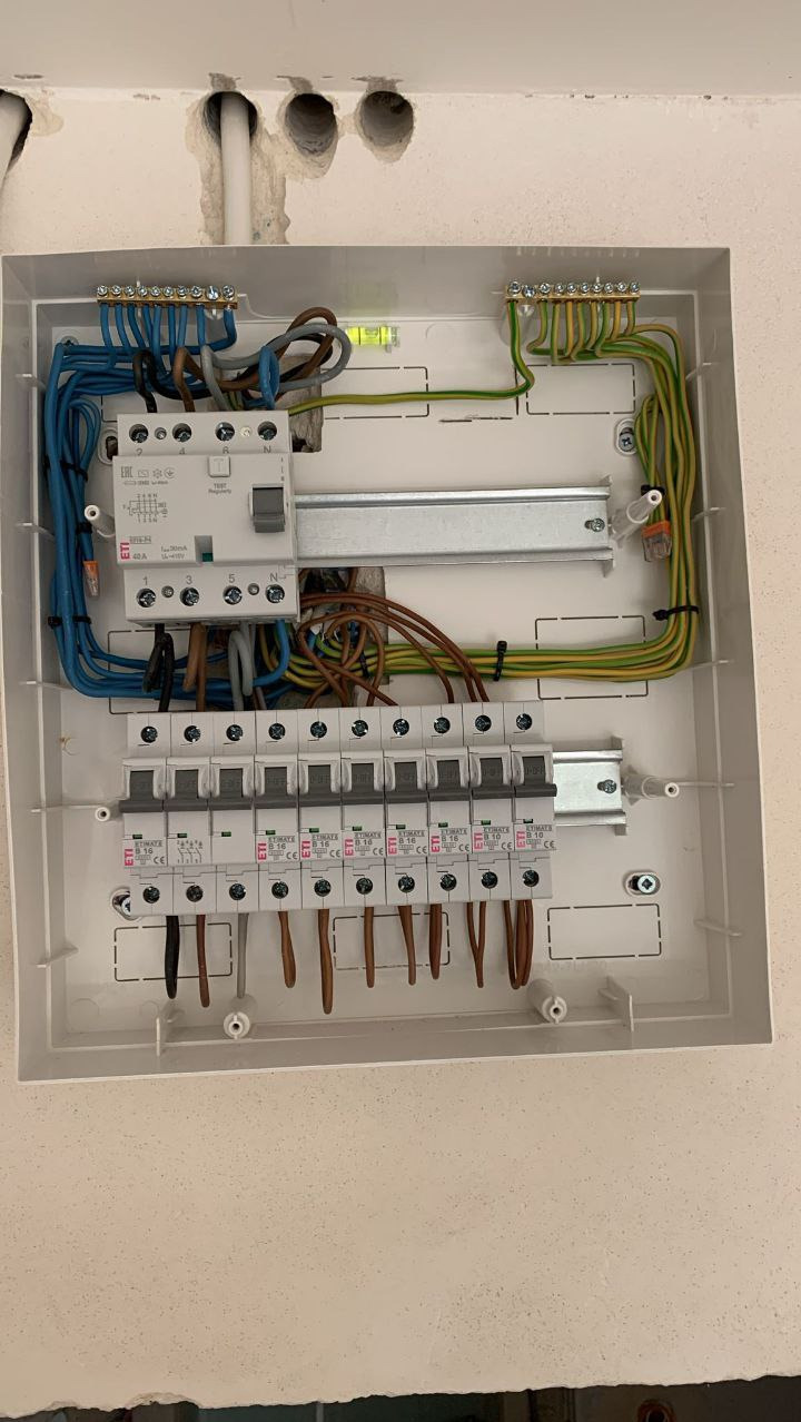 Rozdzielnia elektryczna w białej obudowie z widocznymi bezpiecznikami, przewodami (niebieskie, brązowe, zielono-żółte) i szyną DIN. Instalacja w trakcie montażu.