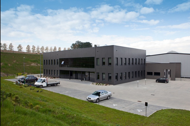 Nowoczesny budynek biurowy o grafitowej elewacji z płyt warstwowych, widok z zewnątrz, plac z kostki brukowej, kilka samochodów na parkingu.