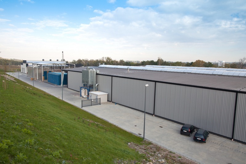 Szeroki kadr hali przemysłowej z szarej płyty warstwowej, z dwoma zaparkowanymi autami. Widok z góry, z zielonym zboczem w lewym dolnym rogu.