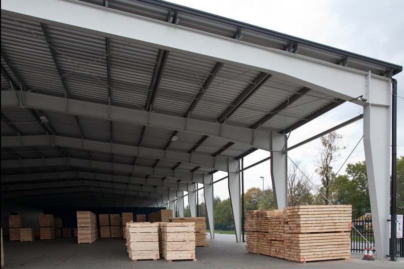 Wnętrze hali stalowej z widoczną konstrukcją dachu i słupami nośnymi. Składowane drewno w paletach. Widok na zewnątrz przez otwartą przestrzeń hali.