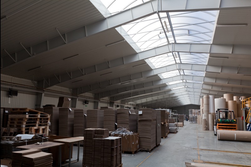 Wnętrze hali produkcyjnej z konstrukcją stalową i świetlikami dachowymi w Wadowicach. Widoczne palety i materiały produkcyjne, co sugeruje etap budowy lub wykończenia.