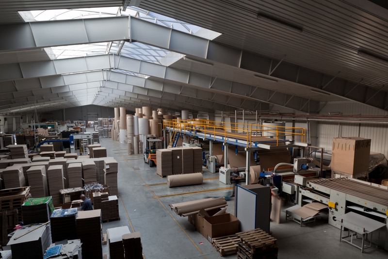 Wnętrze hali produkcyjnej z maszynami, rolami papieru i kartonami. Widoczne świetliki w dachu i wózek widłowy. Perspektywa z góry ukazuje przestrzeń i organizację.