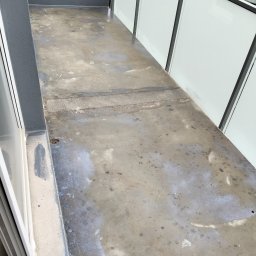 VOLODYMYR MYKHAILYK - Szary, betonowy balkon z widocznymi śladami wilgoci i zabrudzeń. Przezroczyste panele balkonowe z ciemną ramą. Widok na zewnętrzną przestrzeń.