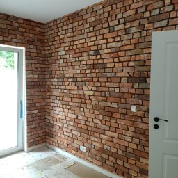 VOLODYMYR MYKHAILYK - Ściana wykończona czerwoną cegłą klinkierową w nowym wnętrzu. Widoczne gniazdka elektryczne, białe drzwi i okno. Na podłodze kartony zabezpieczające posadzkę.