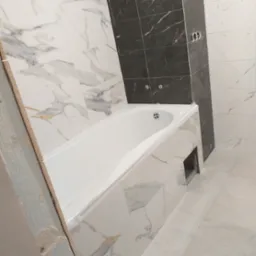 Wykończona łazienka z białą wanną i marmurowymi płytkami, fragment ściany w ciemnych płytkach. Widoczne gniazdka i otwór rewizyjny pod wanną.