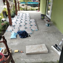 VOLODYMYR MYKHAILYK - Układanie jasnych płytek tarasowych na betonowej wylewce, z widocznymi klipsami poziomującymi i narzędziami. W tle zielona ściana domu, pergola i ogród z basenem.