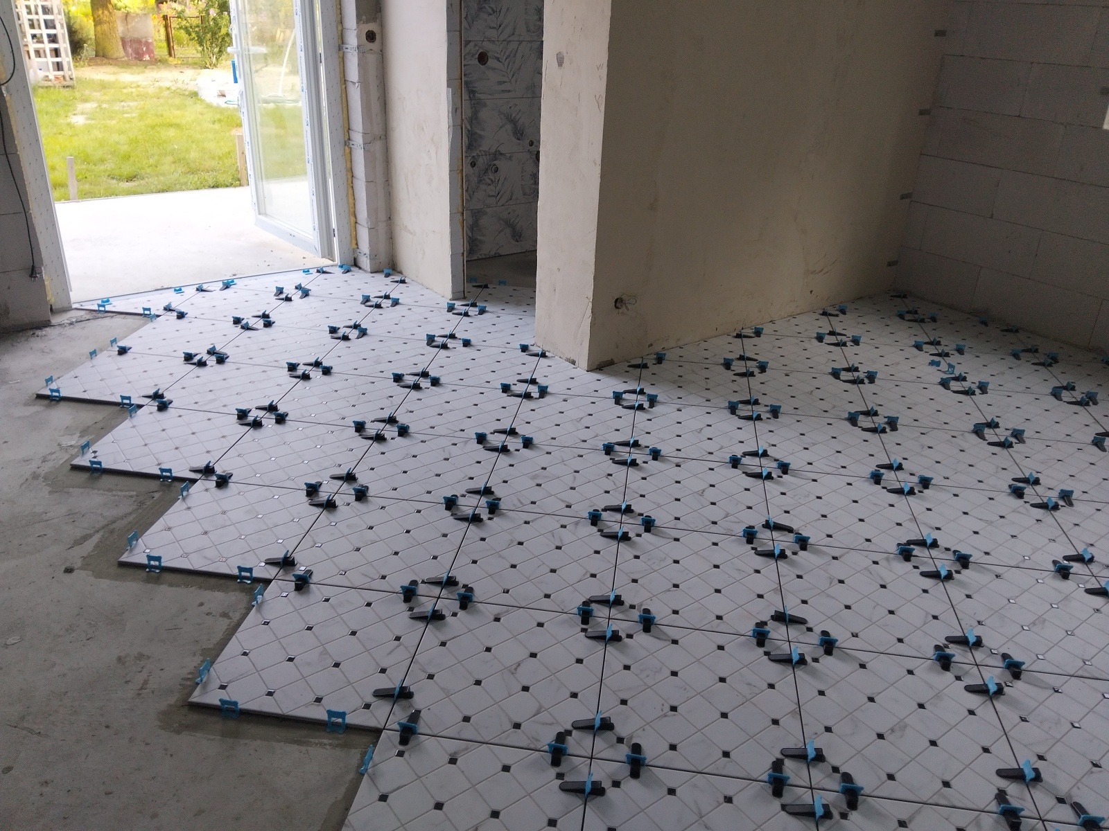 Układanie białych płytek podłogowych z czarnymi akcentami, z użyciem systemu poziomowania płytek. Widoczne niedokończone fragmenty i surowe ściany.