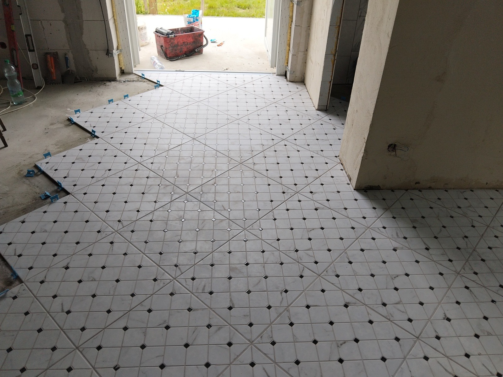 Układanie białych płytek podłogowych z czarnymi akcentami, widoczne niebieskie klipsy poziomujące i narzędzia w tle, wnętrze w trakcie prac wykończeniowych.