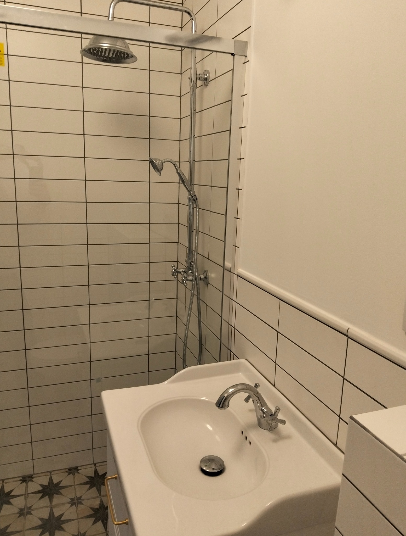 Wykończona łazienka z białymi, poziomymi płytkami, chromowaną armaturą i umywalką w stylu retro na tle wzorzystej podłogi.