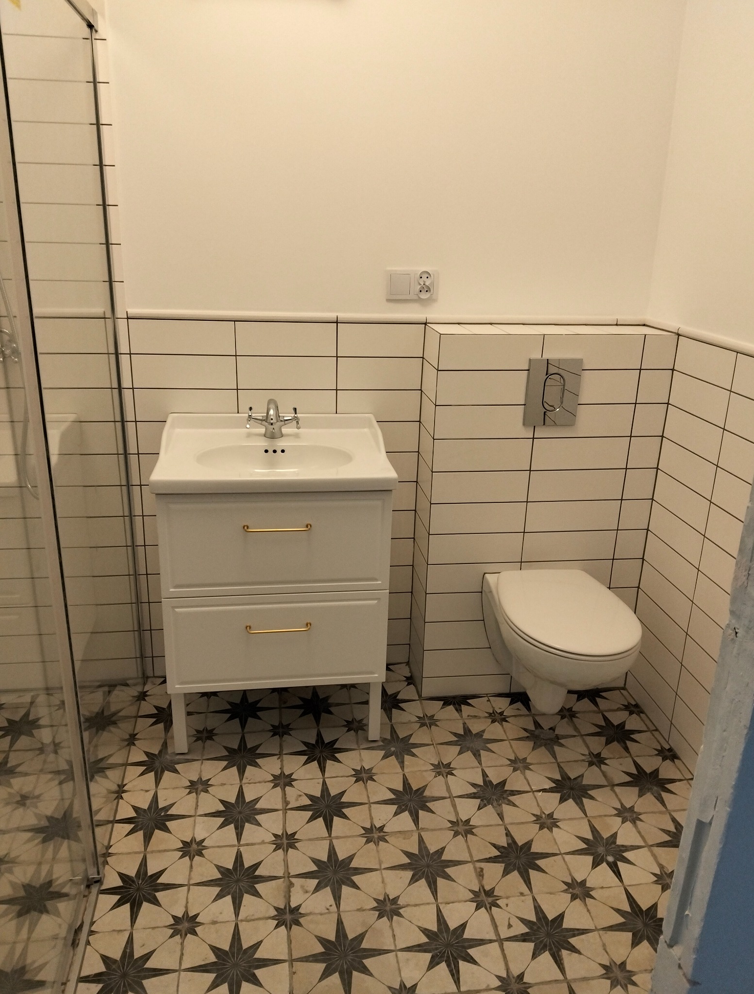 Biała łazienka z umywalką na szafce z złotymi uchwytami, toaletą i wzorzystą podłogą. Ściany wyłożone białymi płytkami z czarną fugą.