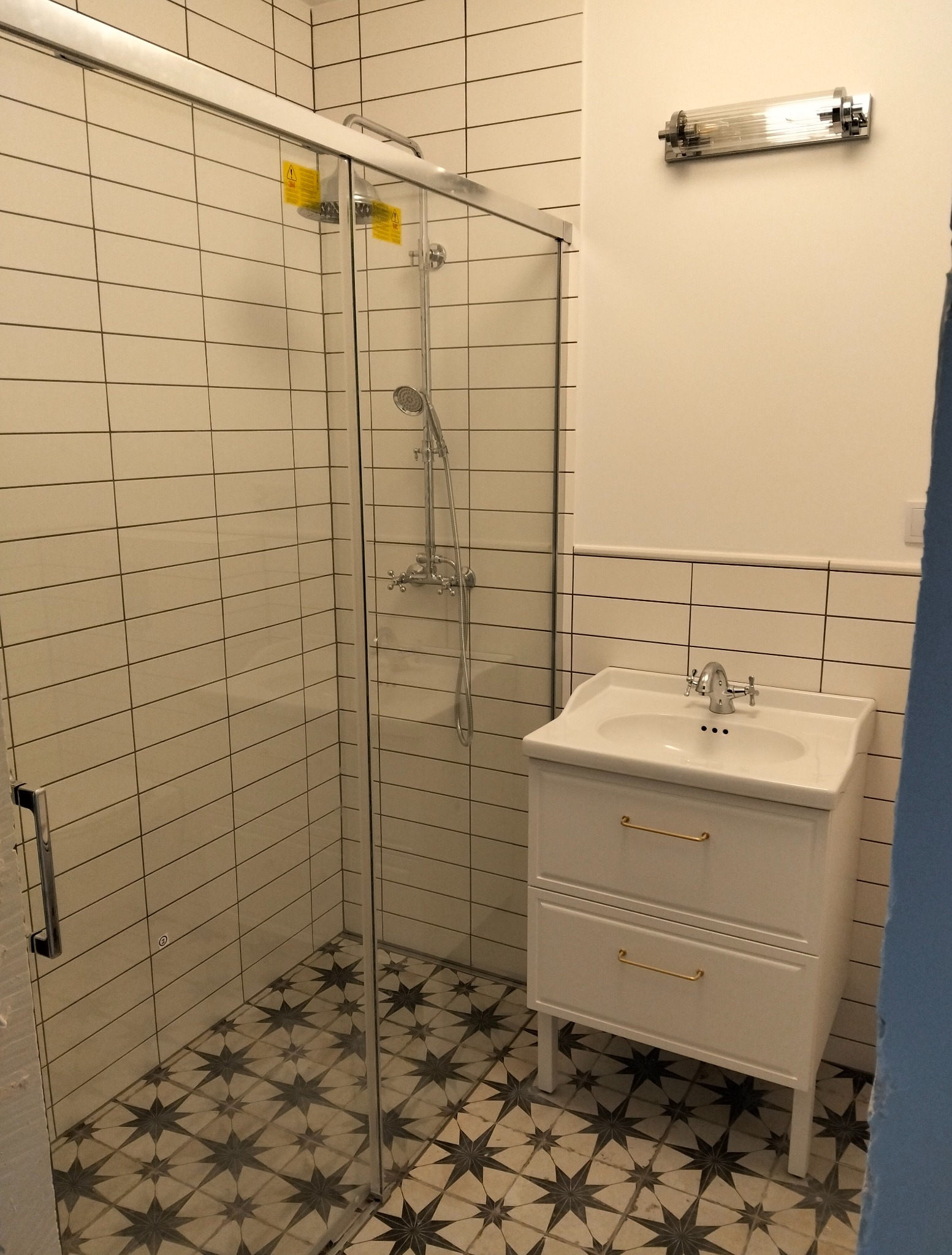 Wykończona łazienka: kabina prysznicowa ze szklanymi drzwiami, umywalka z szafką w stylu retro, podłoga z wzorem w gwiazdy i białe płytki na ścianach.