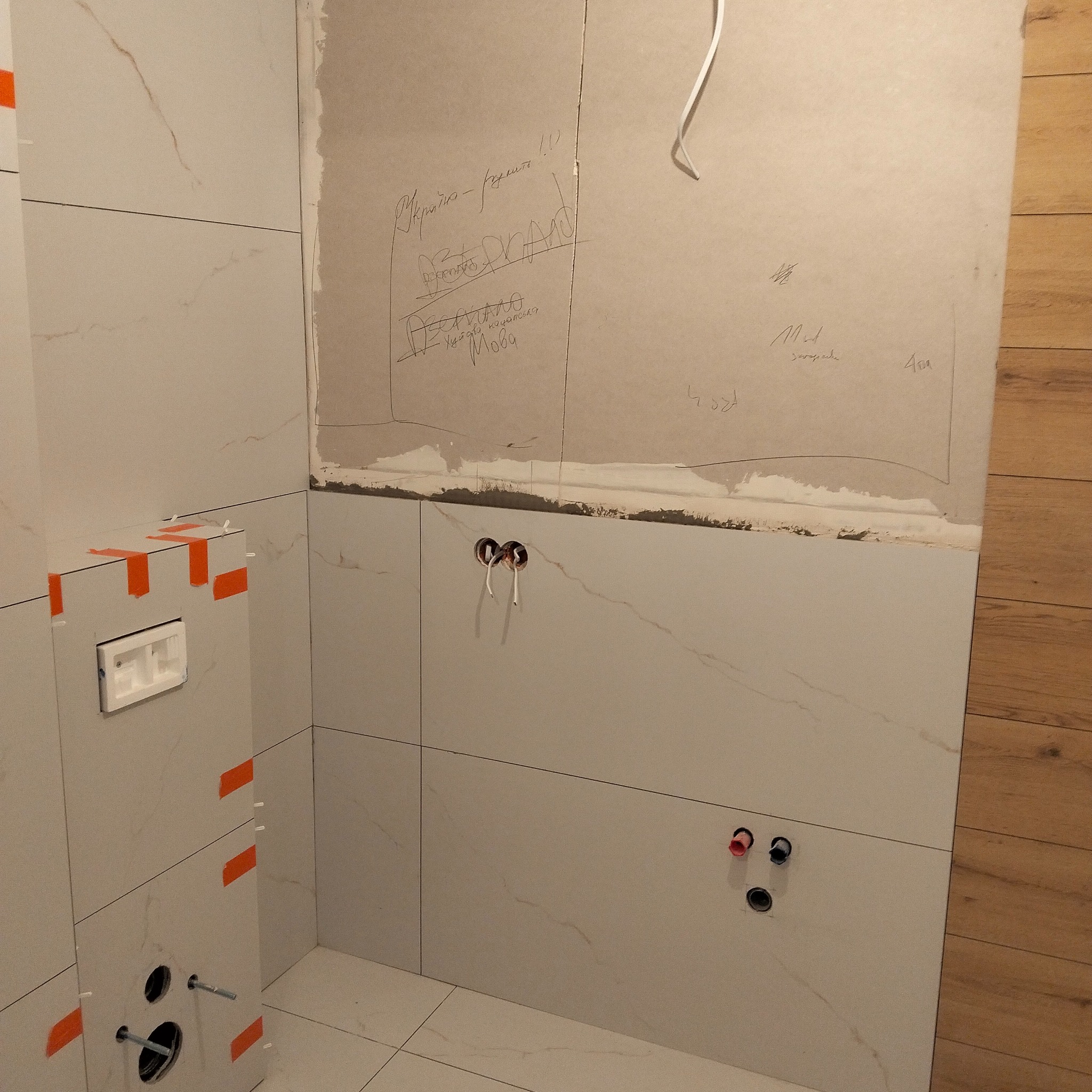 Ściana w trakcie wykańczania w łazience, z widocznymi płytkami imitującymi marmur, instalacją elektryczną i wodną oraz zapiskami na ścianie.