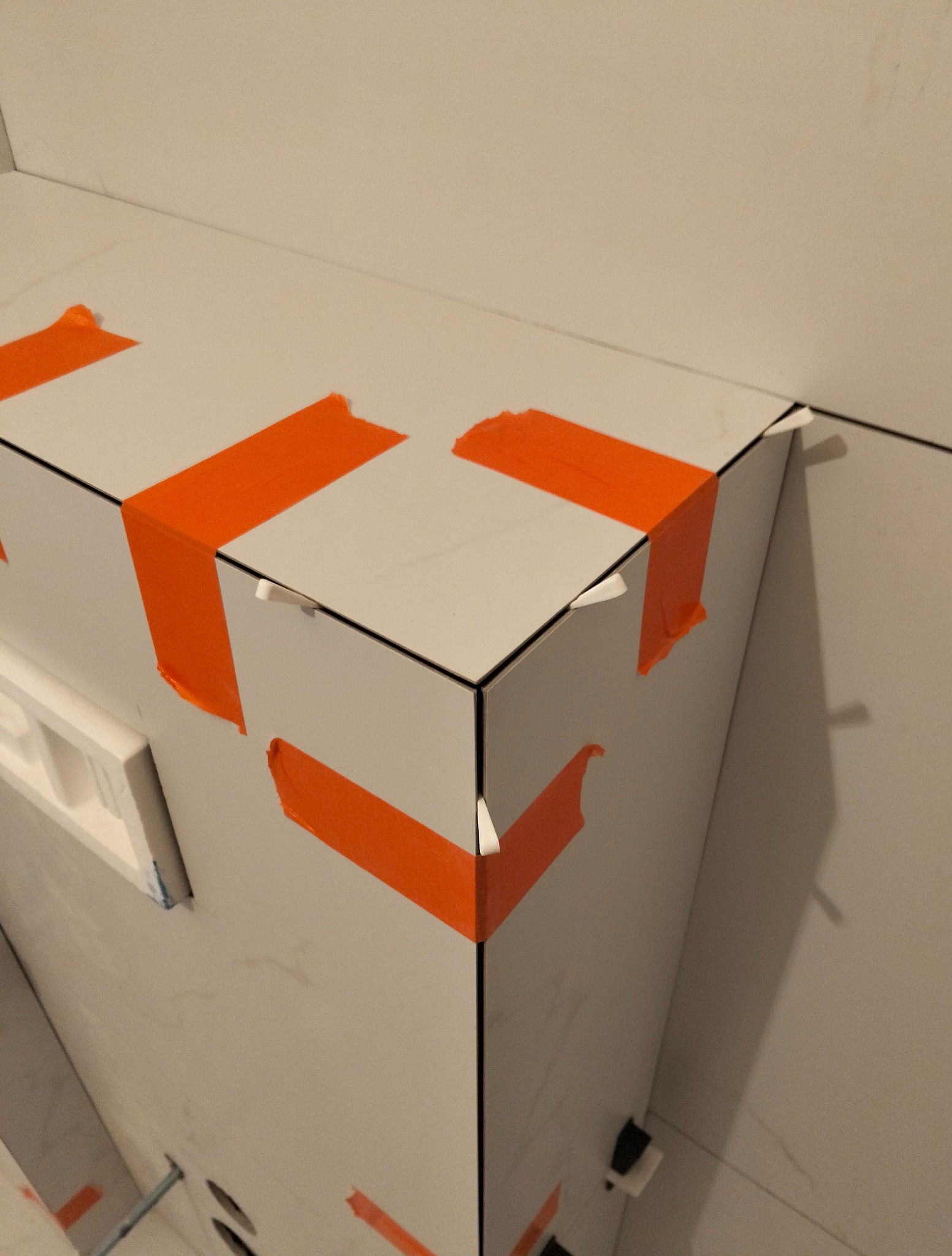 Fragment narożnika wykończonego płytkami, zabezpieczonego pomarańczową taśmą malarską. Widoczne fugi i delikatny wzór marmuru na powierzchni płytek.