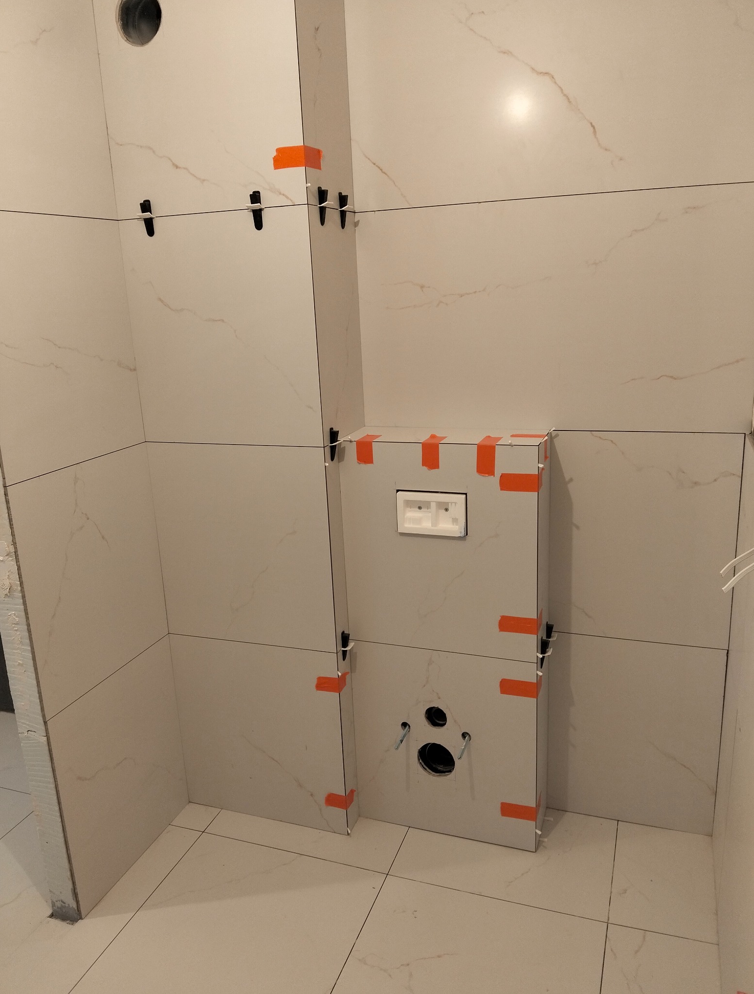 Montaż płytek w łazience, widoczny stelaż podtynkowy z zamontowanymi płytkami, użyte klipsy poziomujące i taśma malarska. Jasne płytki imitujące marmur.