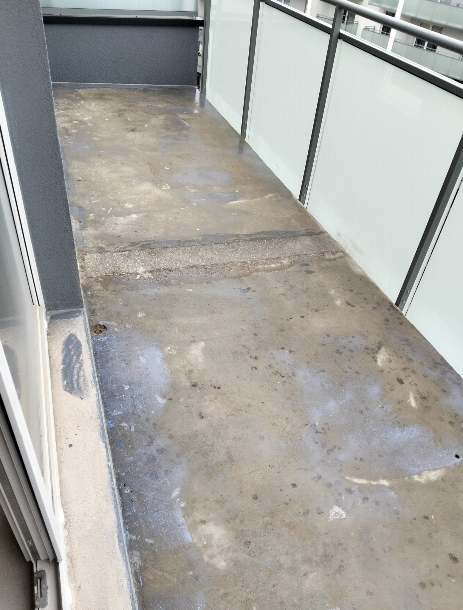 Szary, betonowy balkon z widocznymi śladami wilgoci i zabrudzeń. Przezroczyste panele balkonowe z ciemną ramą. Widok na zewnętrzną przestrzeń.