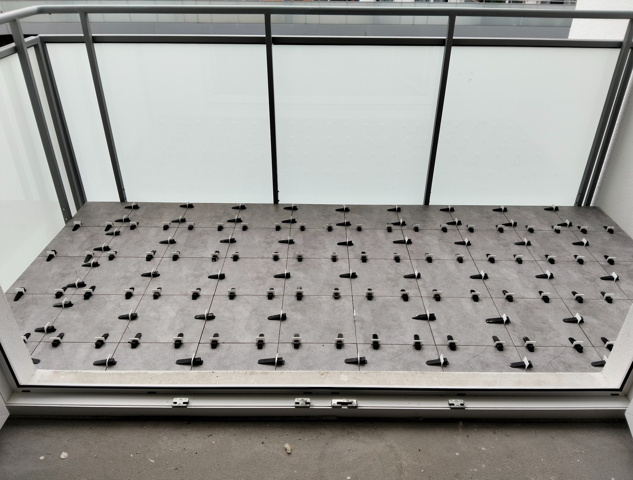 Szare płytki na balkonie w trakcie układania, z użyciem klipsów poziomujących. Widok z góry, częściowo widoczna balustrada i szyba.