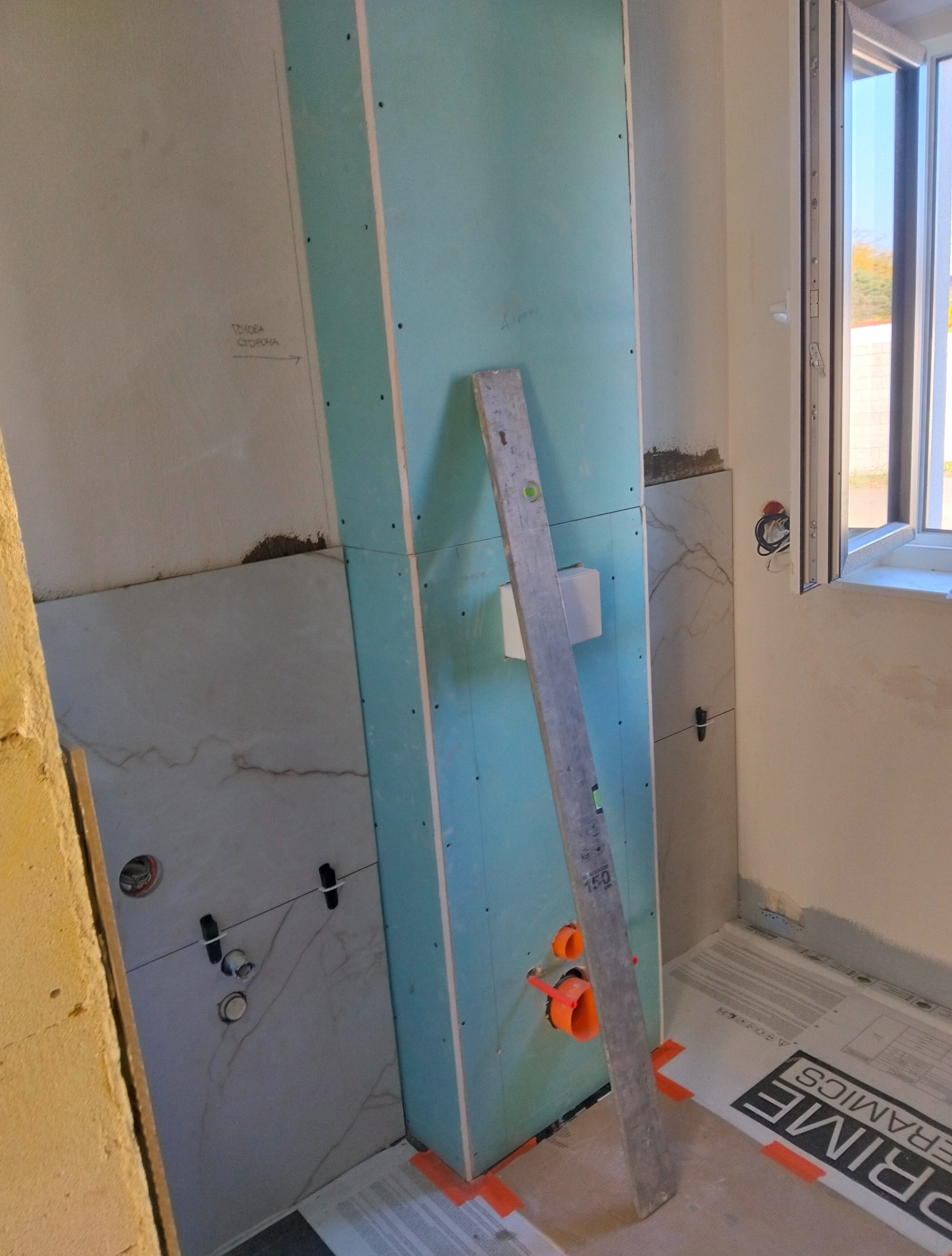 Montaż stelaża podtynkowego w łazience z widoczną instalacją hydrauliczną i poziomnicą. Częściowo położone płytki imitujące marmur.