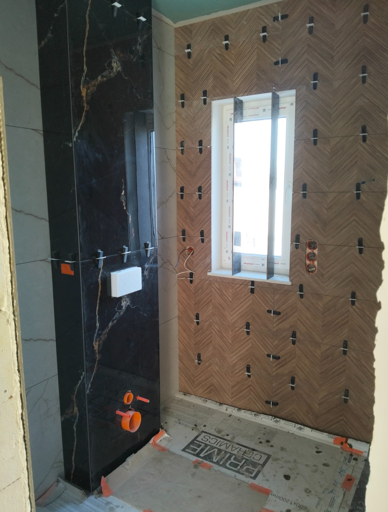 Łazienka w trakcie wykończenia: montaż płytek w jodełkę wokół okna, obok ściana z czarnych płytek imitujących marmur. Widoczne elementy instalacji i podkład zabezpieczający.