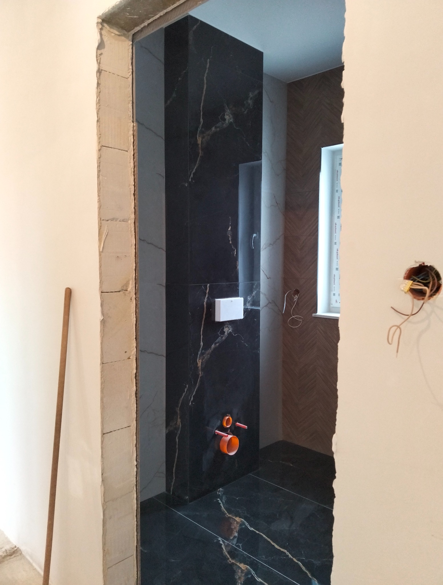 Wykończona łazienka z czarnymi płytkami imitującymi marmur i widoczną instalacją. Ujęcie przez surową futrynę drzwi.