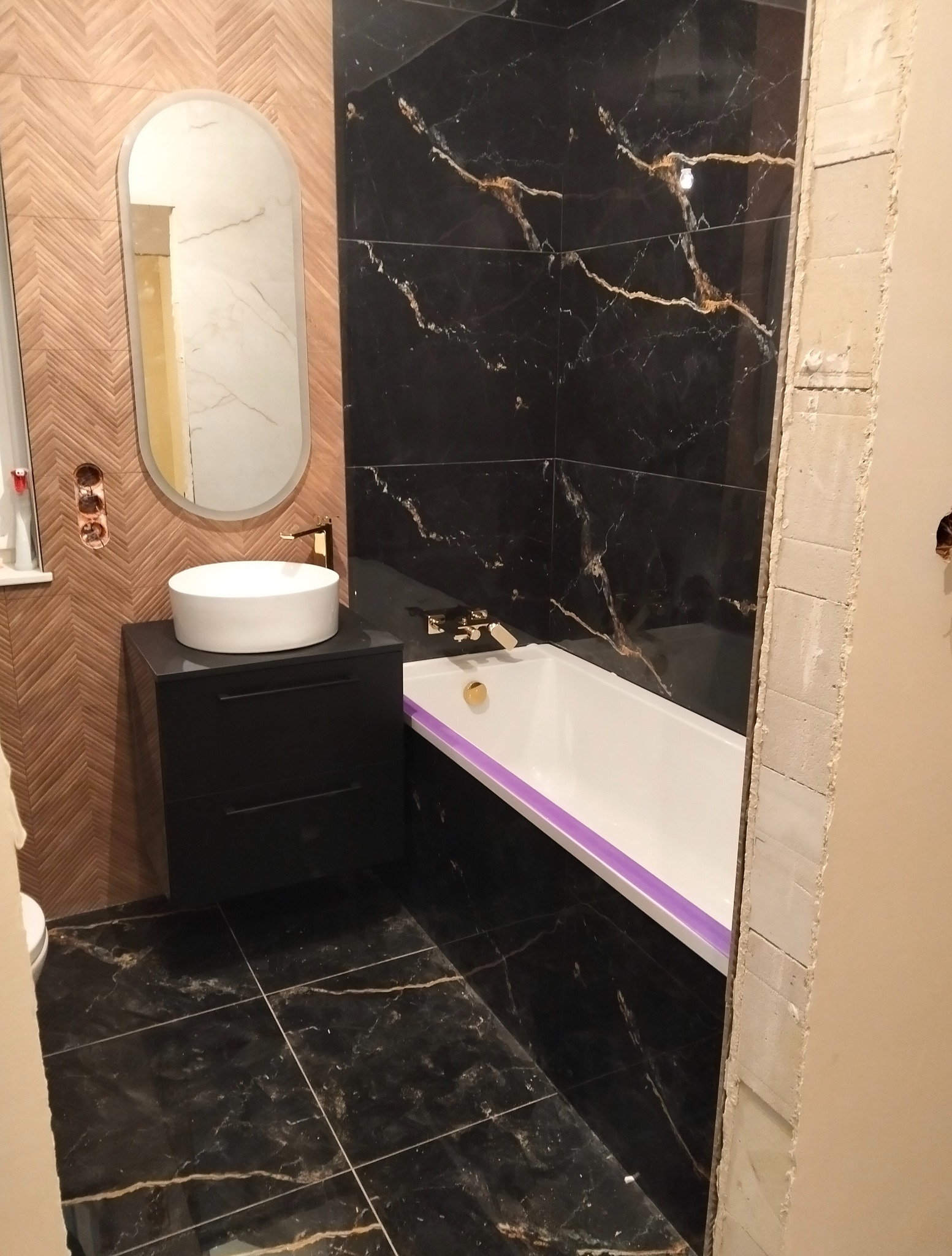 Wykończona łazienka z czarnymi płytkami imitującymi marmur, drewnianą ścianą w jodełkę, owalnym lustrem i białą umywalką nablatową. Złota armatura.