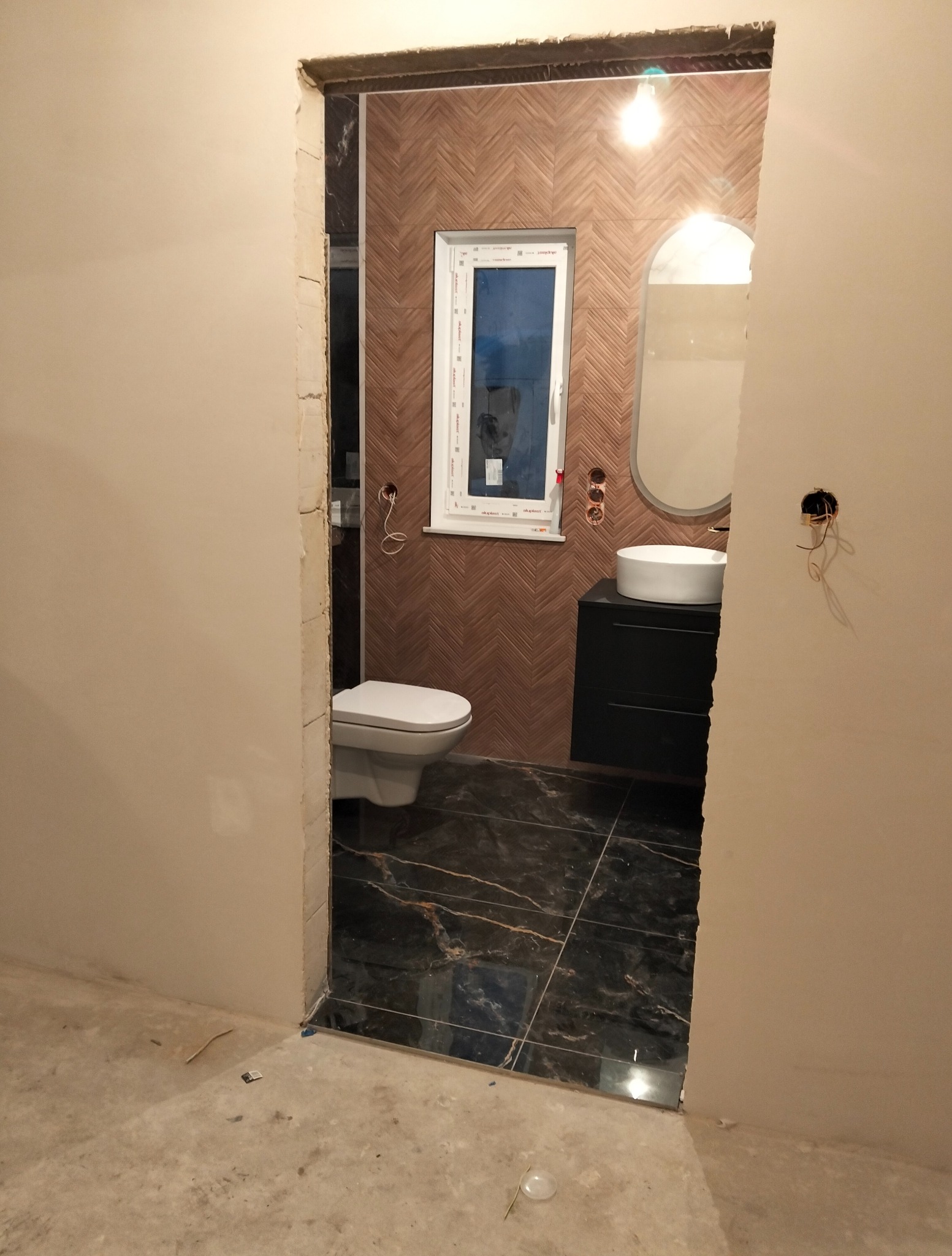 Wykończona łazienka z brązowymi płytkami ułożonymi w jodełkę, czarną podłogą z żyłami i owalnym lustrem nad czarną szafką z białą umywalką.