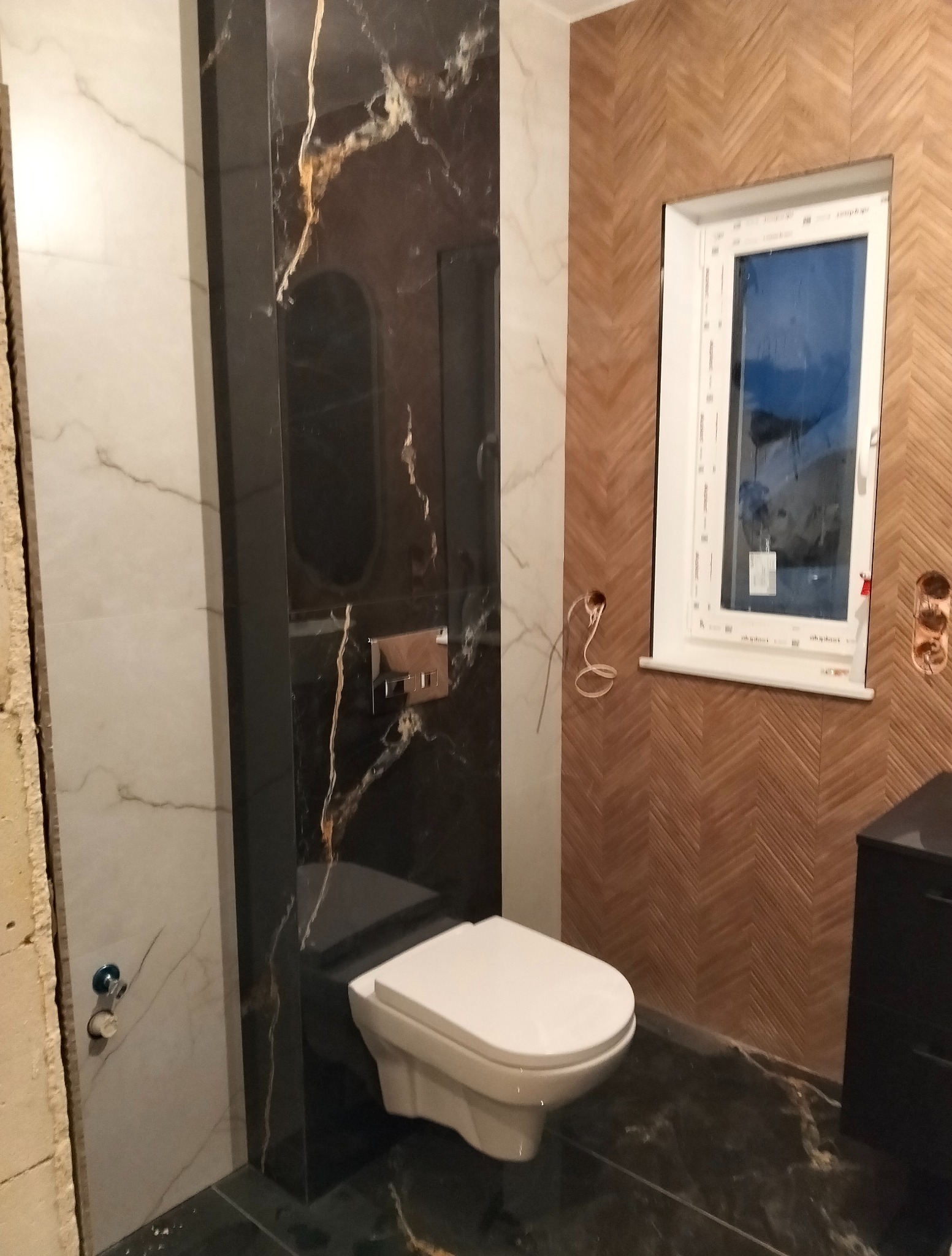 Elegancka toaleta z marmurową obudową, drewnianą ścianą w jodełkę i nowoczesną armaturą. Widoczna czarna szafka i okno z białymi ramami.