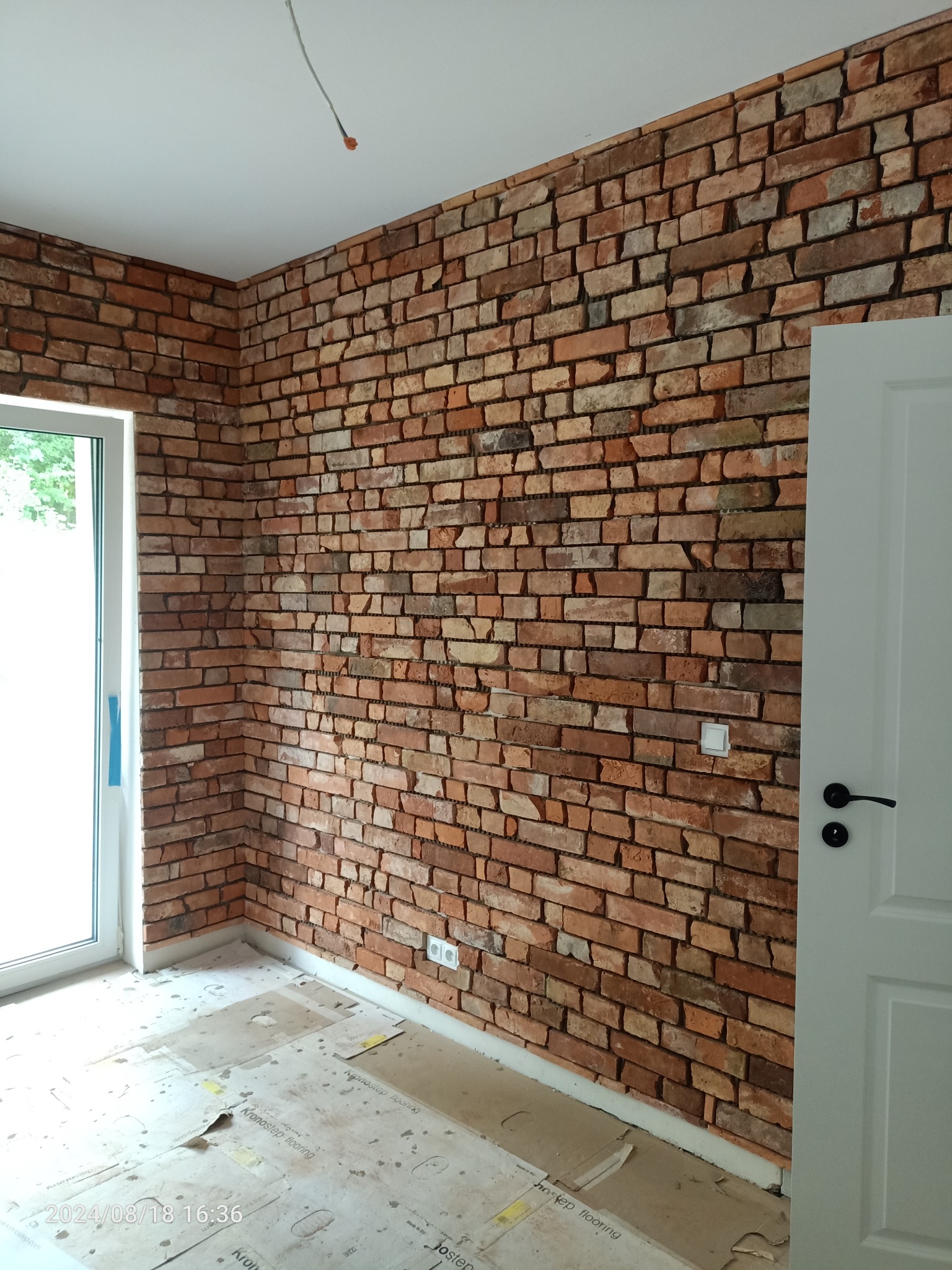 Ściana wykończona czerwoną cegłą klinkierową w nowym wnętrzu. Widoczne gniazdka elektryczne, białe drzwi i okno. Na podłodze kartony zabezpieczające posadzkę.