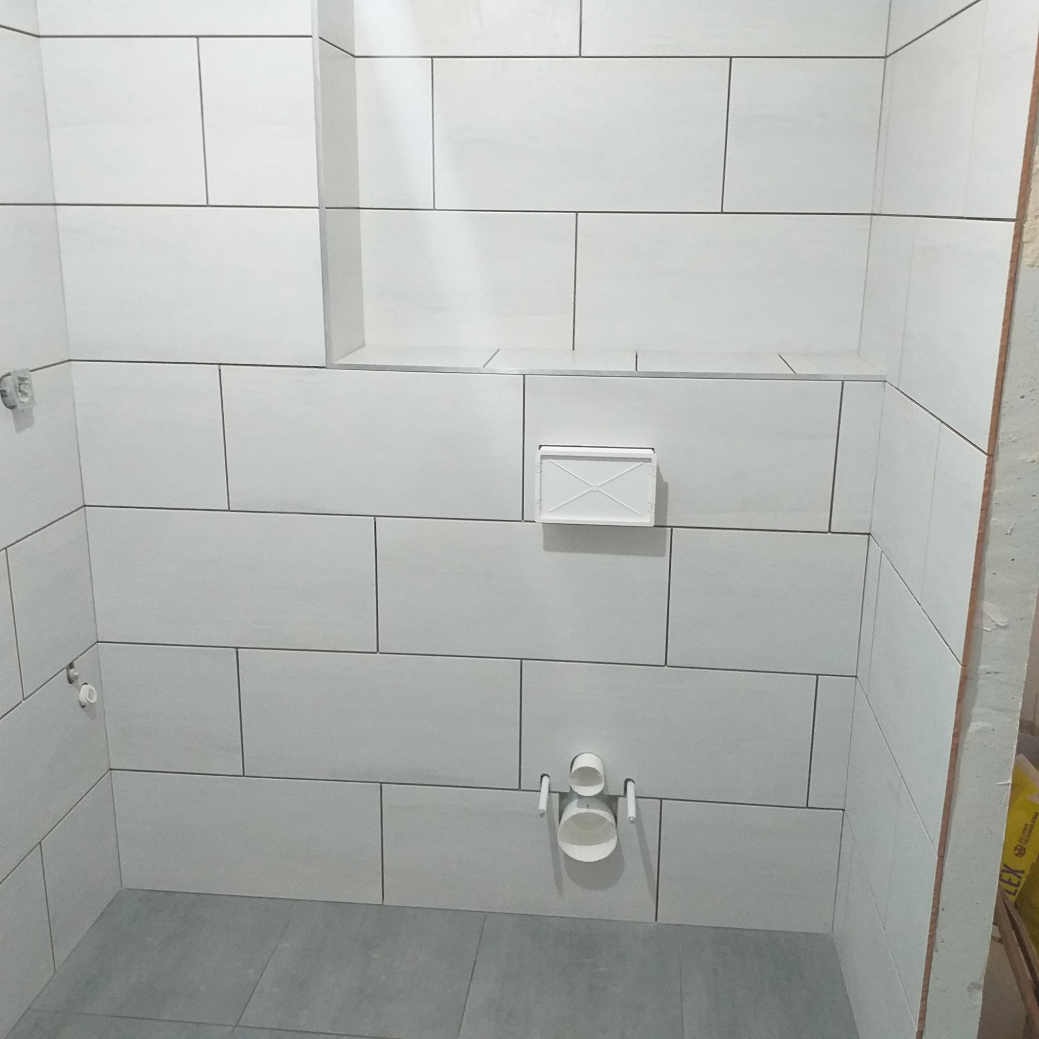 Wykończona ściana łazienki z białymi, prostokątnymi płytkami i szarą podłogą. Widoczny fragment instalacji wodnej i zabudowa stelaża WC.