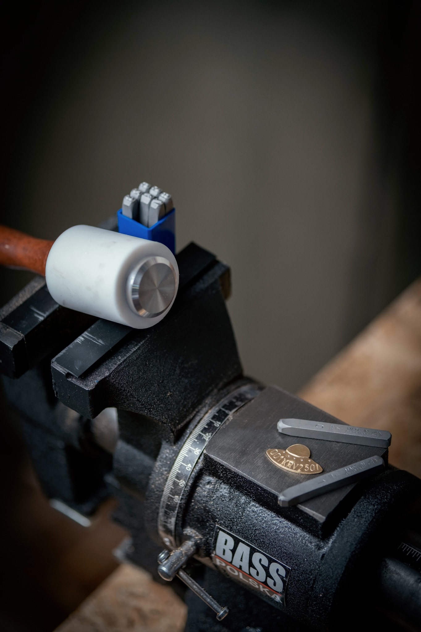 Metalowa blaszka z wygrawerowanym napisem w imadle, obok stemple i młotek. Narzędzia ślusarskie w warsztacie. Rękodzieło.