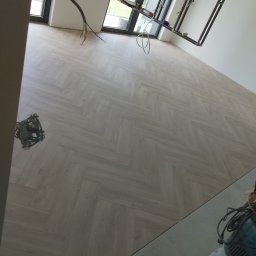 Arkadiusz Szwarc - Jasna podłoga ułożona w jodełkę francuską w nowym mieszkaniu, z widocznymi narzędziami i instalacją elektryczną, w trakcie wykańczania, blisko okna.