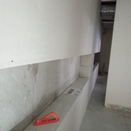 Arkadiusz Szwarc - Narożnik z płyty karton-gips w trakcie montażu, widoczne narzędzia: poziomica i śrubokręt. Surowy stan wykończenia wnętrza, widoczne elementy konstrukcyjne.