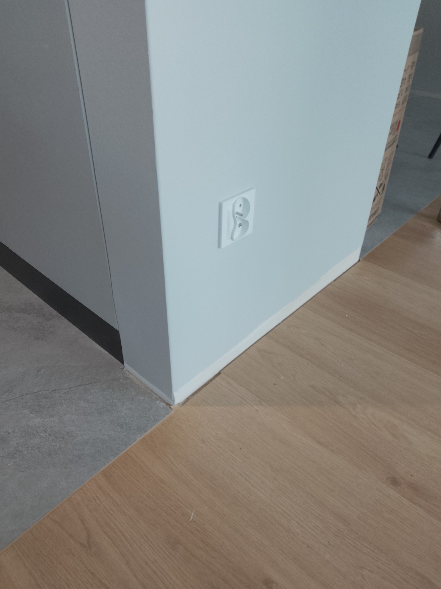 Styk drewnianej podłogi z szarą płytką u dołu ściany z gniazdkiem. Widoczne listwy przypodłogowe. Ujęcie z bliska na wykończenie podłogi i ścian.