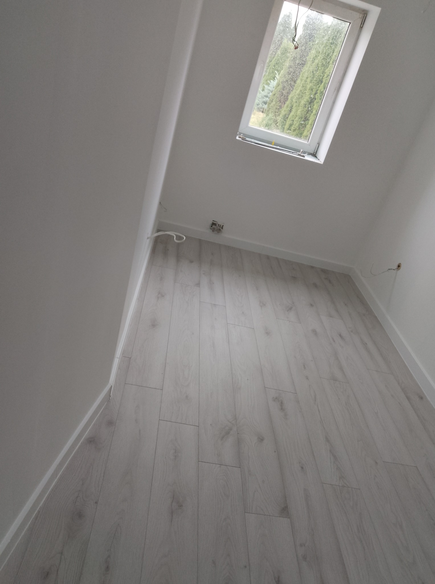 Jasne panele podłogowe w pokoju na poddaszu, widok z góry. Ujęcie obejmuje okno dachowe i fragmenty białych ścian. Minimalistyczny wystrój.