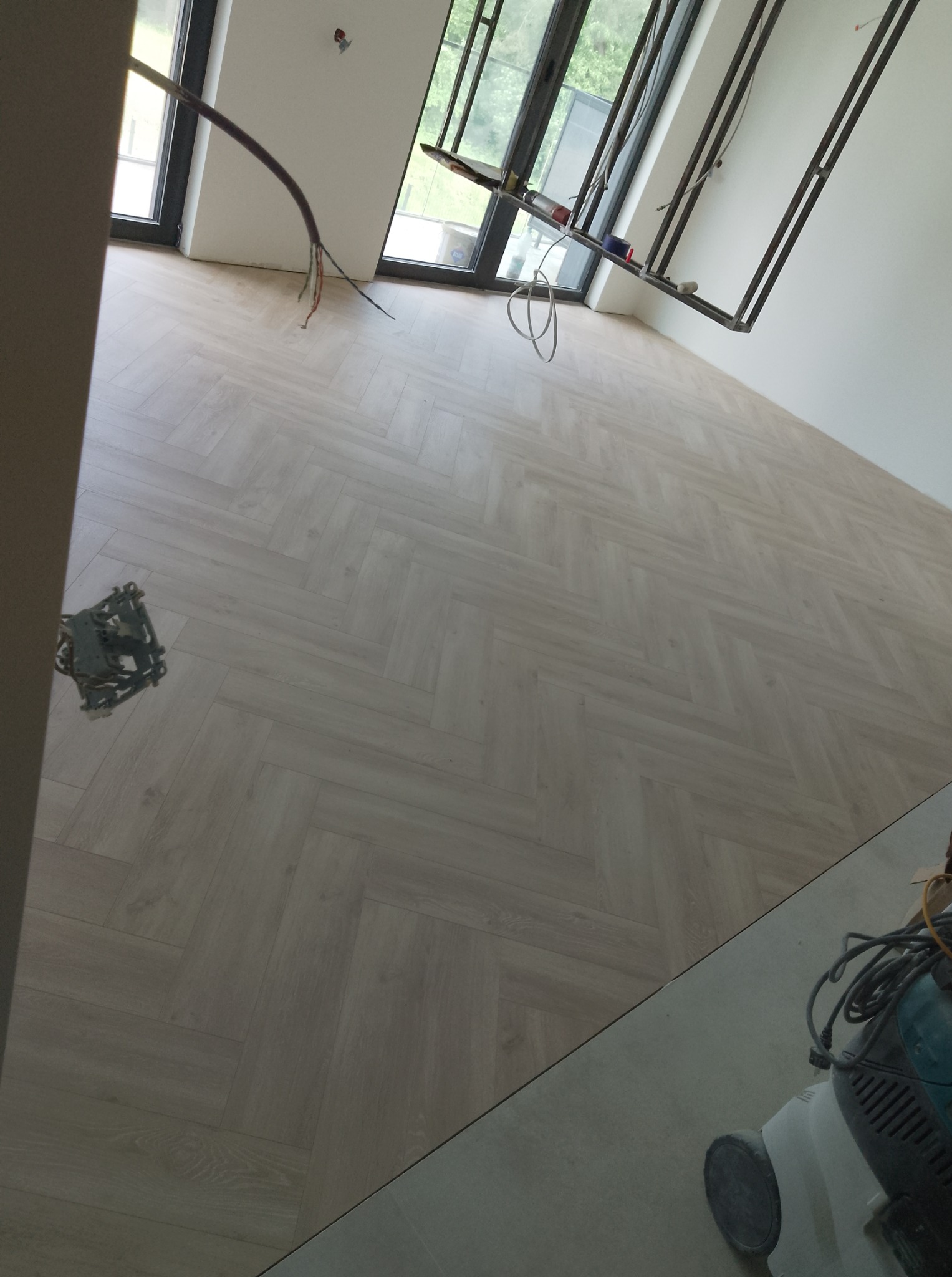 Jasna podłoga ułożona w jodełkę francuską w nowym mieszkaniu, z widocznymi narzędziami i instalacją elektryczną, w trakcie wykańczania, blisko okna.