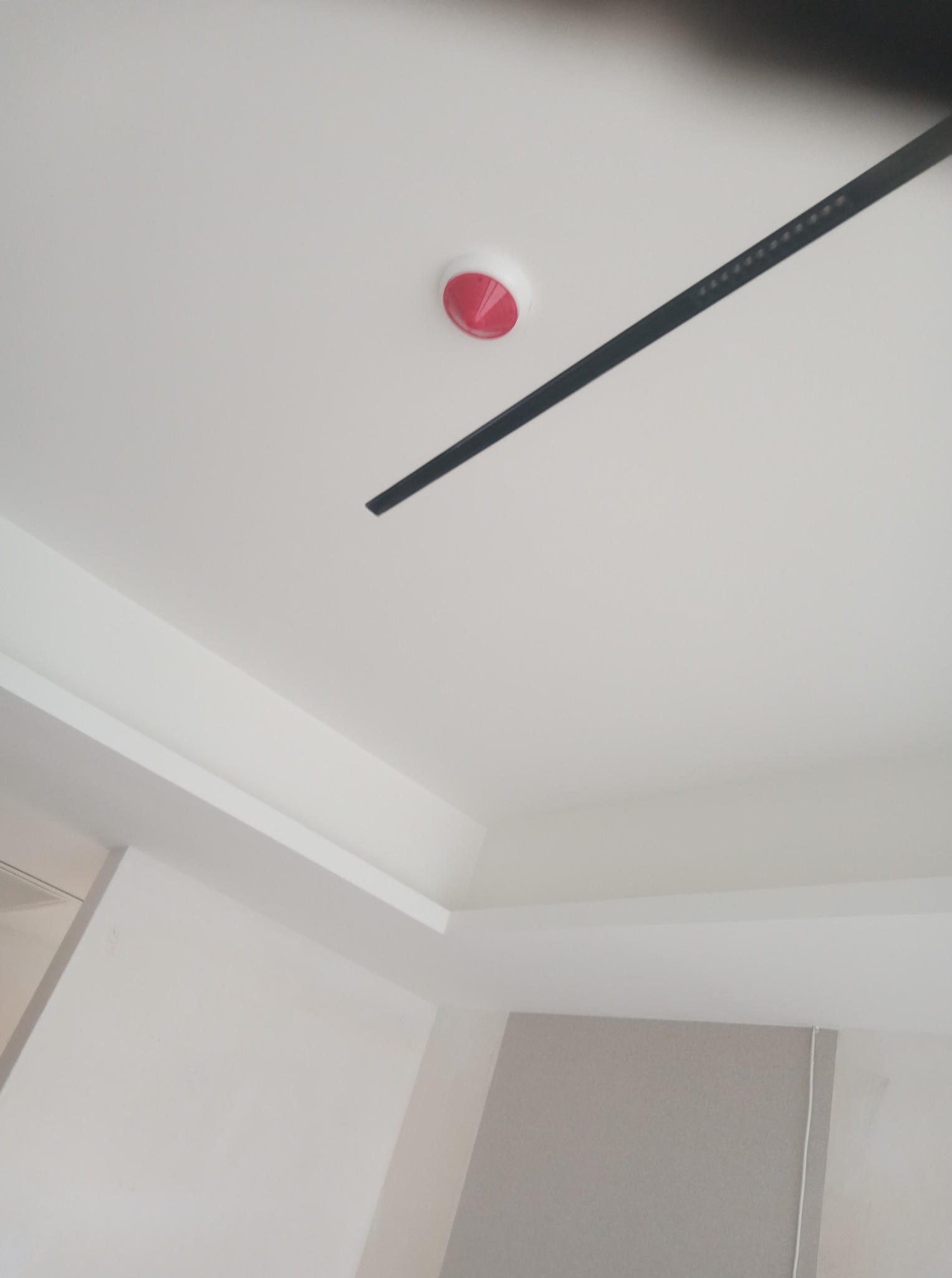 Biały sufit z czarną listwą oświetleniową i czujnikiem dymu z czerwoną obwódką. Fragment ściany w tle, wnętrze po remoncie.