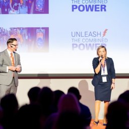 Aleksandra Paluch - Prelegenci na scenie podczas konferencji z ekranem wyświetlającym prezentację, widownia w tle. Kobieta z mikrofonem i mężczyzna w garniturze.