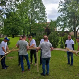 Zesp&oacute;ł mężczyzn w plenerze łączy drewniane elementy konstrukcji podczas integracji firmowej, na tle zieleni i budynk&oacute;w.