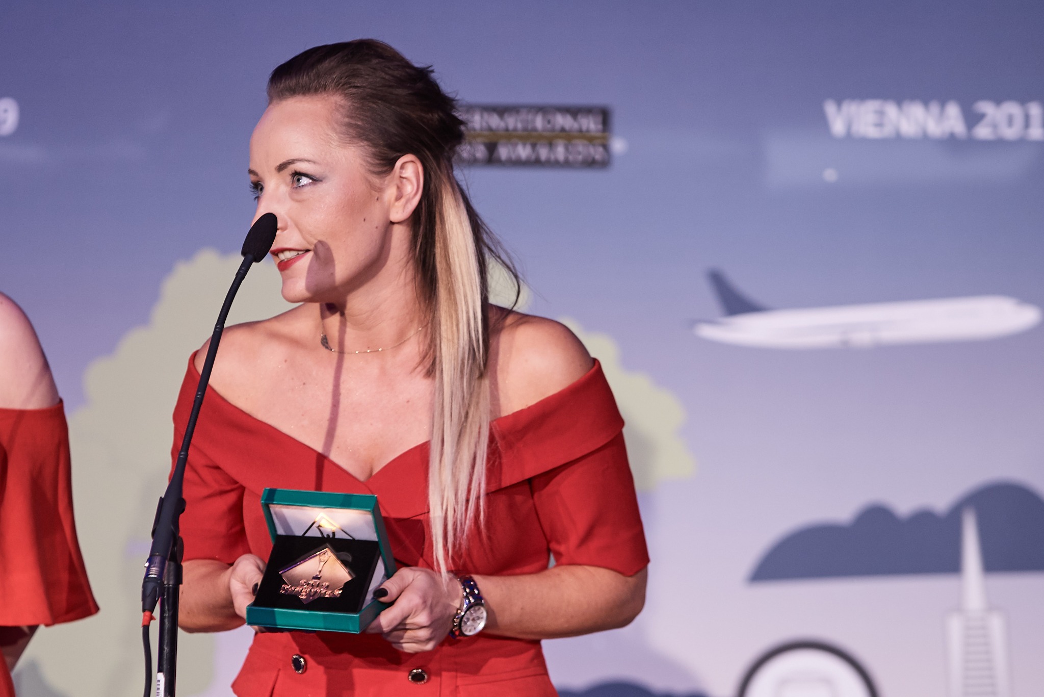 Kobieta w czerwonej sukience odbiera nagrodę podczas konferencji w Wiedniu, trzymając otwarte pudełko z odznaczeniem, mikrofon przed nią.