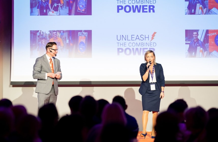 Prelegenci na scenie podczas konferencji z ekranem wyświetlającym prezentację, widownia w tle. Kobieta z mikrofonem i mężczyzna w garniturze.