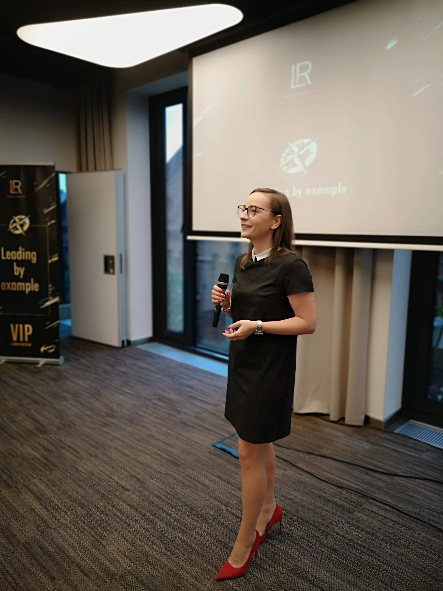 Kobieta w okularach i czerwonych szpilkach przemawia z mikrofonem na tle ekranu projekcyjnego z logo, podczas szkolenia biznesowego.