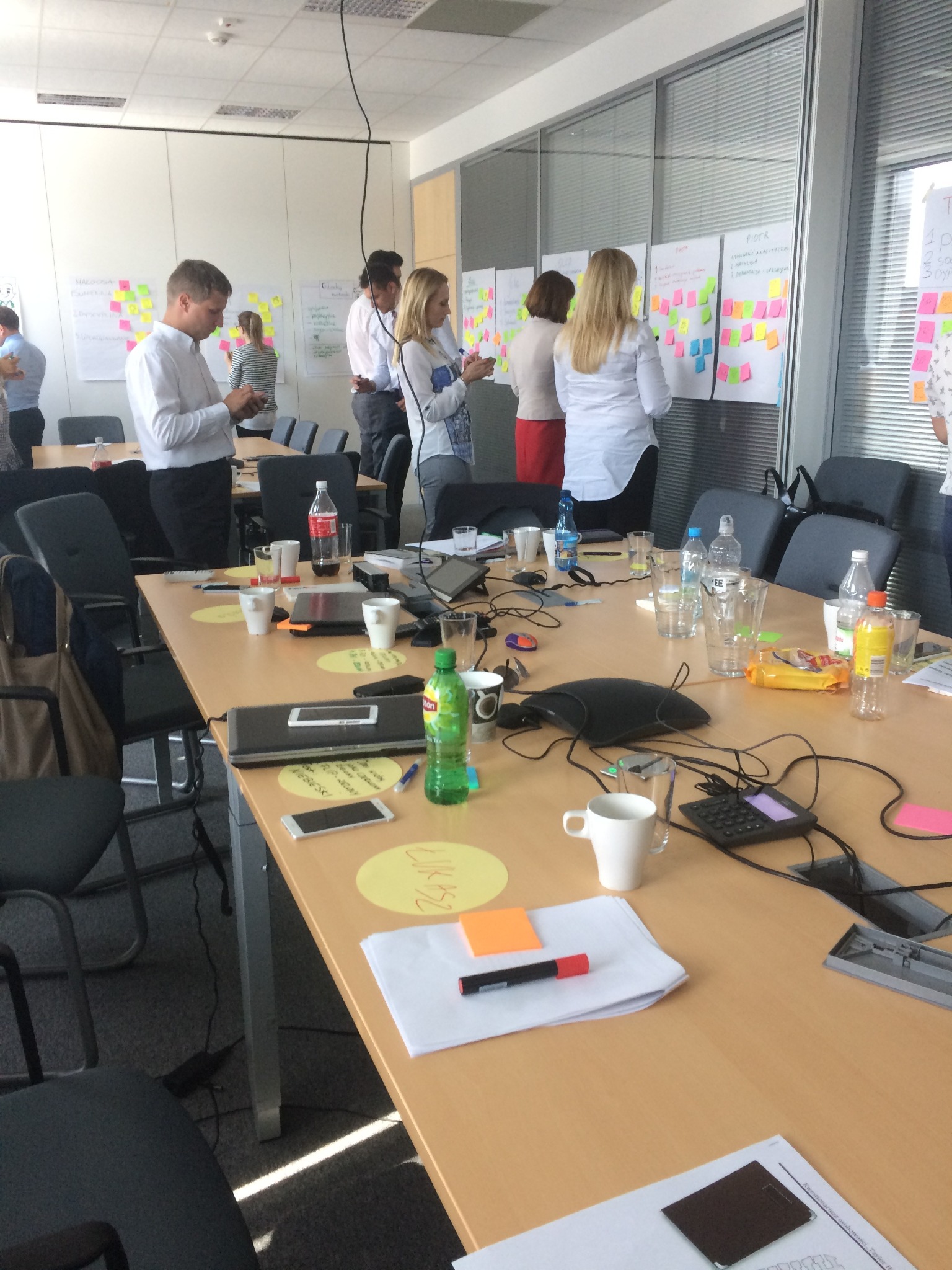 Sala konferencyjna podczas szkolenia: uczestnicy analizują tablice z notatkami. Na stole widoczne materiały szkoleniowe, napoje i przekąski. Jasne, naturalne światło.