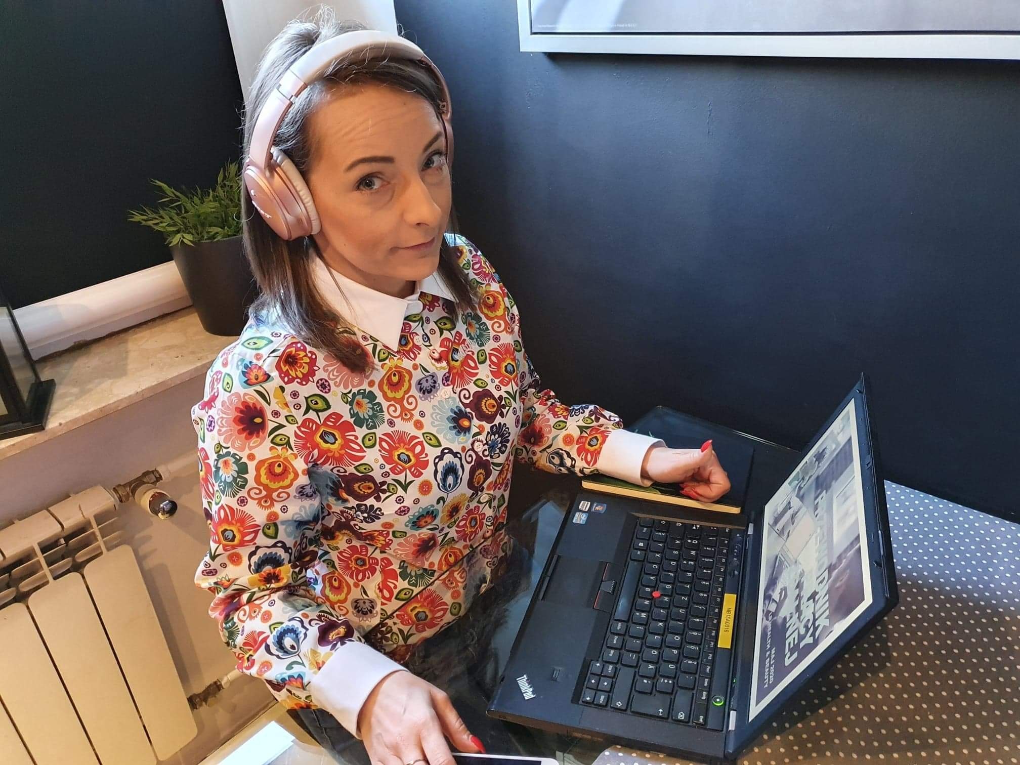 Kobieta w słuchawkach, w kwiecistej bluzce, pracuje na laptopie przy biurku. Widoczny notatnik i telefon. W tle ściana i roślina doniczkowa.