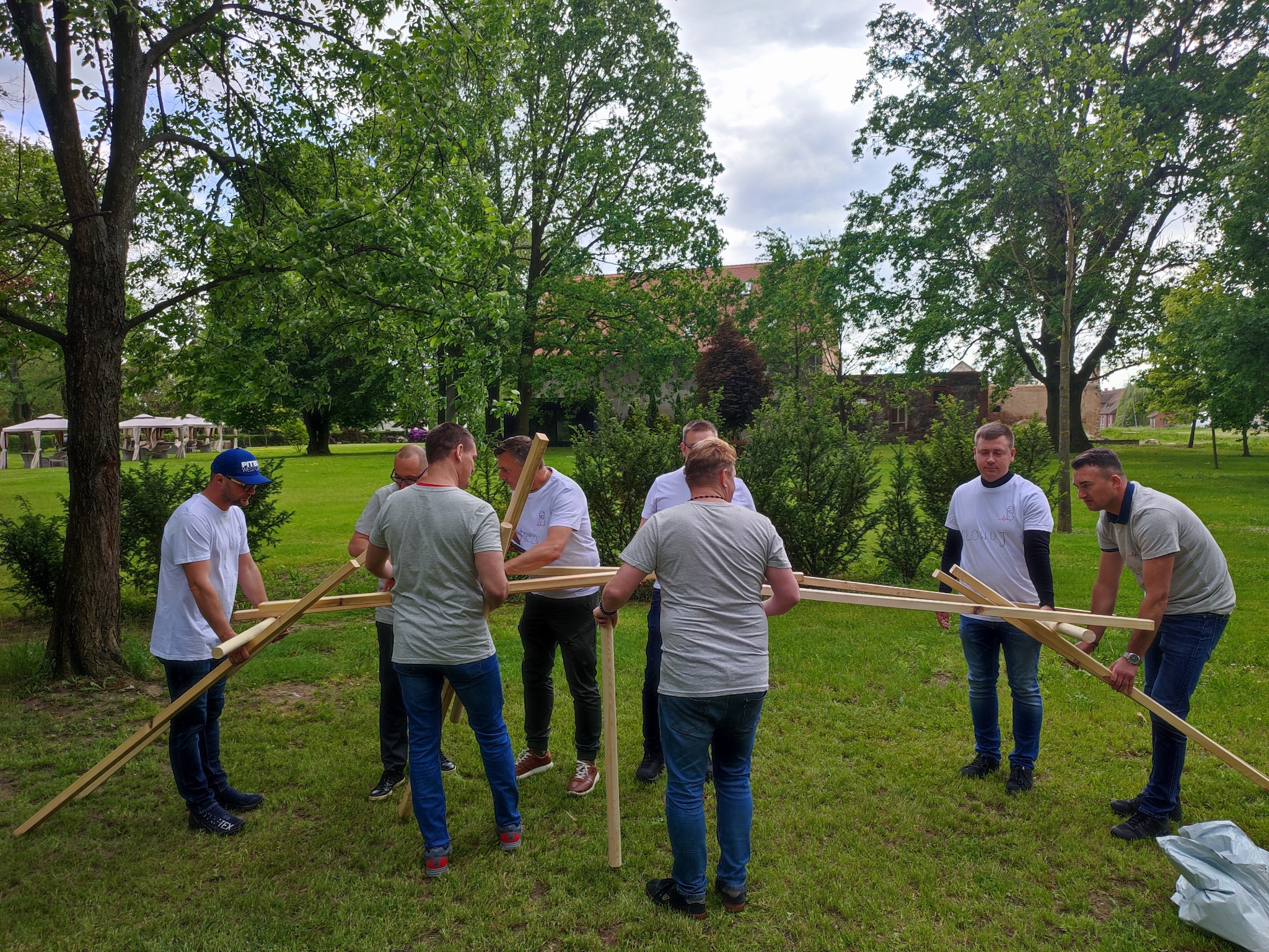 Zespół mężczyzn w plenerze łączy drewniane elementy konstrukcji podczas integracji firmowej, na tle zieleni i budynków.